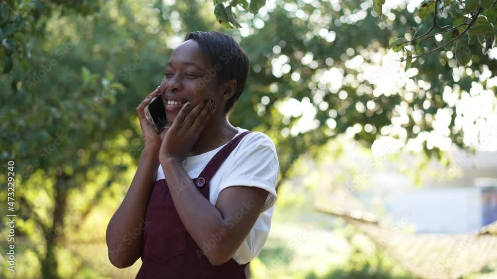 Portrait of slim confident beautiful African American woman talking on phone standing in sunset sunrays outdoors. Smiling young lady enjoying leisure in summer park in slow motion Portrait of slim confident beautiful African American woman talking on phone standing in sunset sunrays outdoors. Smiling young lady enjoying leisure in summer park in slow motion