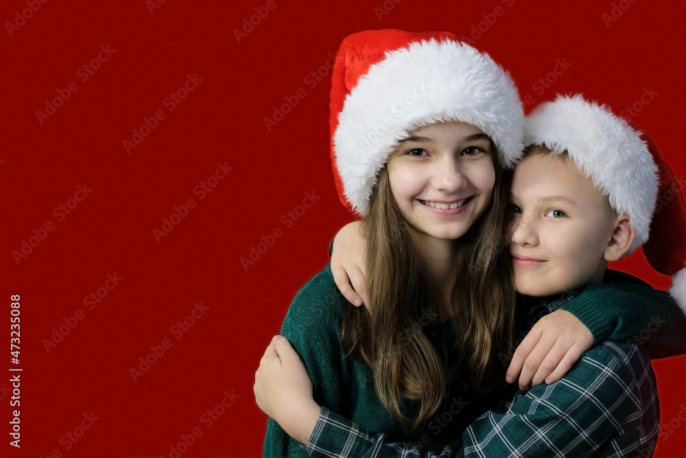 A girl and a boy in Santa Claus hats and green clothes on a red background hug and smile. They look into the camera. Copy space A girl and a boy in Santa Claus hats and green clothes on a red background hug and smile. They look into the camera. Copy space