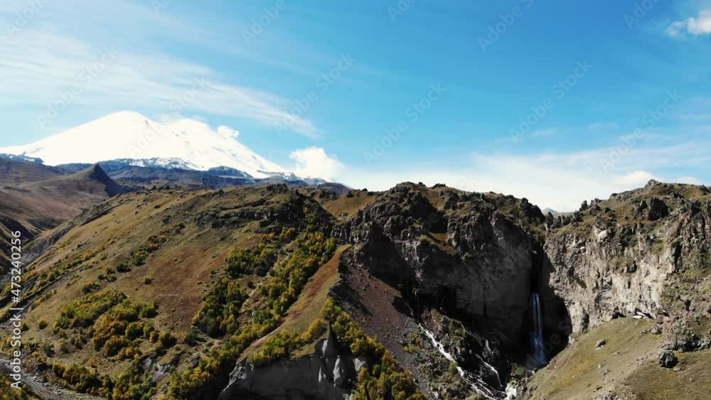 Aerial view of mountain waterfall and Mount Elbrus. View of snowy white cloudless peak of Mount Elbrus. Stormy river flows down from top of mountain into gorge and breaks on the ground Aerial view of mountain waterfall and Mount Elbrus. View of snowy white cloudless peak of Mount Elbrus. Stormy river flows down from top of mountain into gorge and breaks on the ground