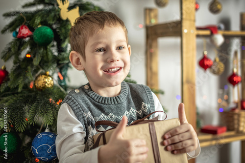 Happy little boy is standing next to a Christmas tree in a decorated festive room and holding a gift box in his hands. Concept of a holiday atmosphere and New Year mood