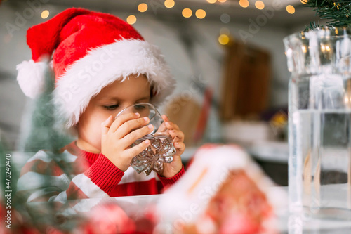 Happy funny toddler boy in a red Santa hat drinking filtered water from a glass in the kitchen. Holidays, health concept.