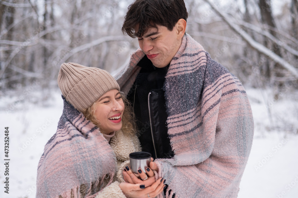 A young couple walks in the park in winter. The guy and the girl are drinking a warming drink outdoors. A young couple walks in the park in winter. The guy and the girl are drinking a warming drink outdoors.