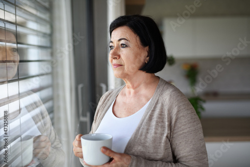 Sad senior woman standing and looking out through window indoors at home.