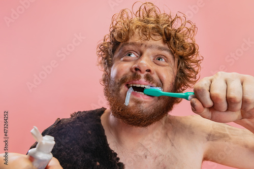 Cropped portrait of funny man in character of neanderthal brushing teeth with toothpaste isolated over pink background photo