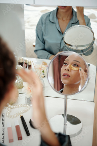 Young woman looking at mirror when applying moisturizing undereye patches