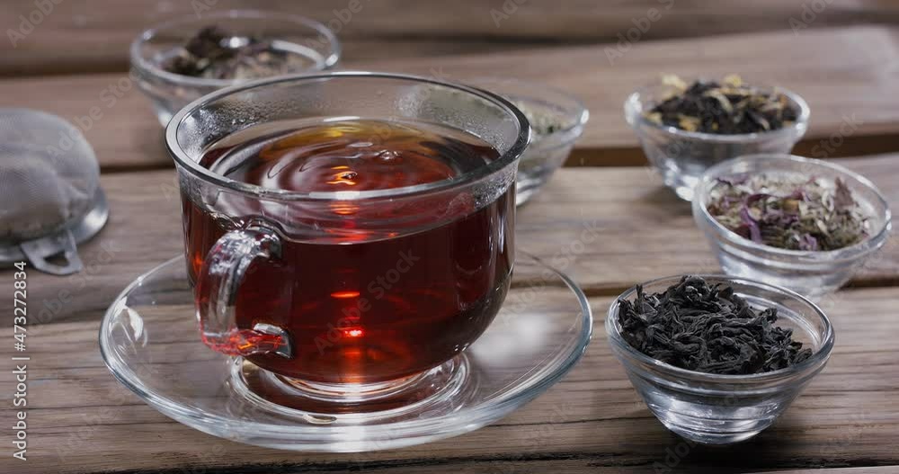 Drop slowly falls into a glass cup of tea. Around there are small bowls with different types of leaf tea. Vintage wooden table. Drop slowly falls into a glass cup of tea. Around there are small bowls with different types of leaf tea. Vintage wooden table.