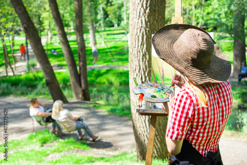 The woman artist paints a picture in a spring park with a resting people.