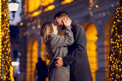 Christmas people hugging and cuddling on the street on Christmas eve. A young happy romantic couple standing on the street surrounded by Christmas light, hugging and cuddling on Christmas eve. © dusanpetkovic1