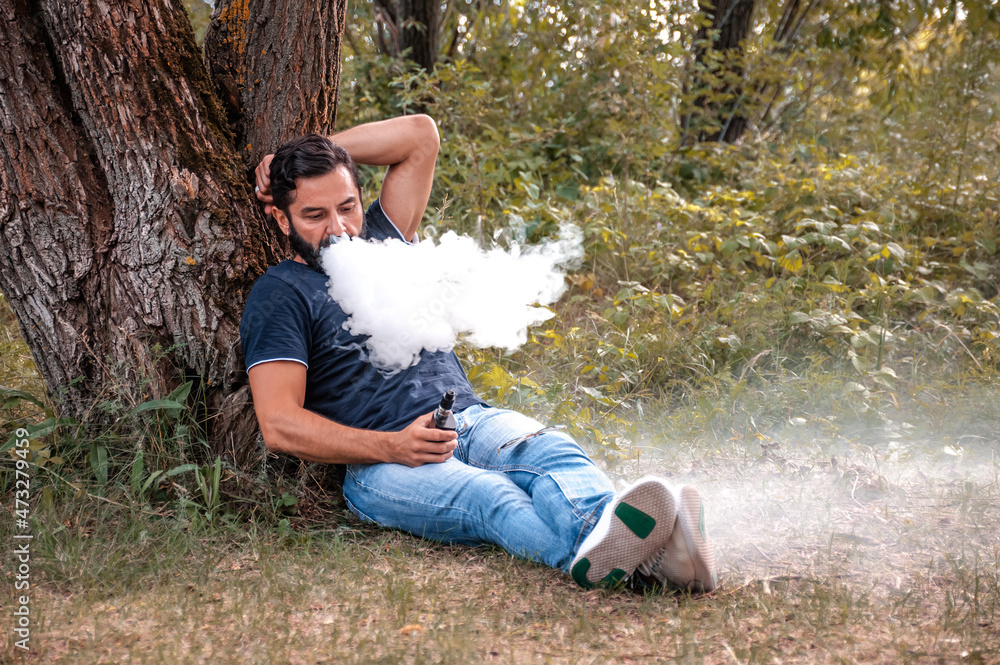 Stylish bearded smoker blows up a couple an e-cigarette on the forest floor. Electronic cigarette. Stylish bearded smoker blows up a couple an e-cigarette on the forest floor. Electronic cigarette.