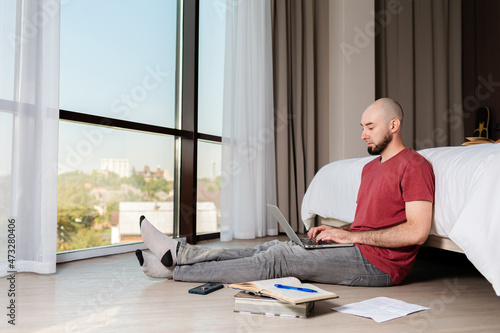 A young Caucasian man sits on the floor and types on a laptop. Spacious room with large windows. The concept of online courses and online learning