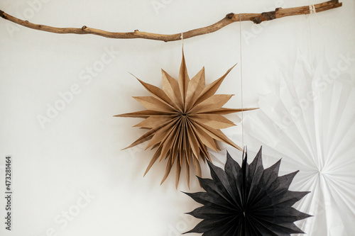 Stylish Christmas stars hanging on wooden branch on white wall. Modern festive scandinavian decor in room. Simple sweden craft, black and white paper stars. Winter holiday preparation.