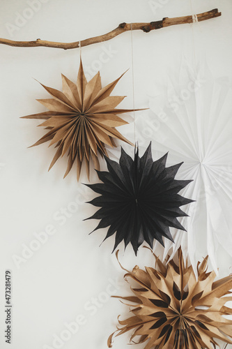 Stylish Christmas stars hanging from wooden branch on white wall. Modern festive scandinavian decor in room. Simple black, craft, white sweden paper stars. Winter holiday preparation.