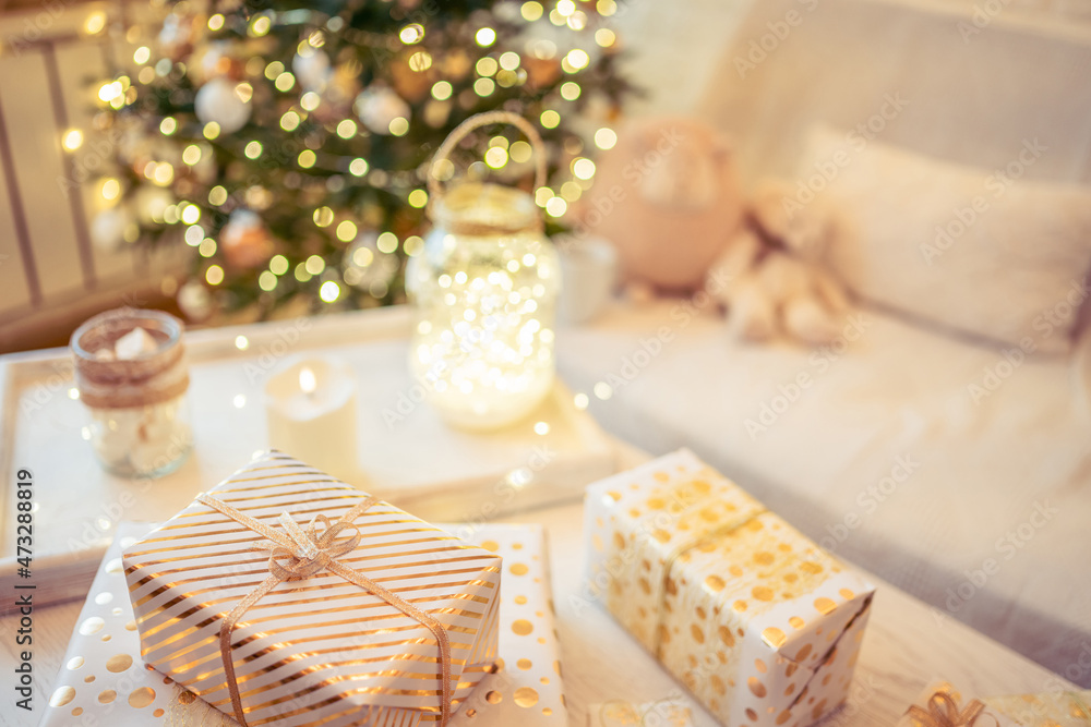 A lot of packing handmade gift boxes lying on the table near Christmas tree in the midst of golden lights, glowing garland, candle. Soft focus