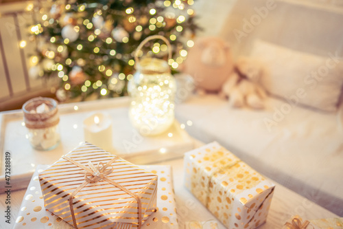 A lot of packing handmade gift boxes lying on the table near Christmas tree in the midst of golden lights  glowing garland  candle. Soft focus