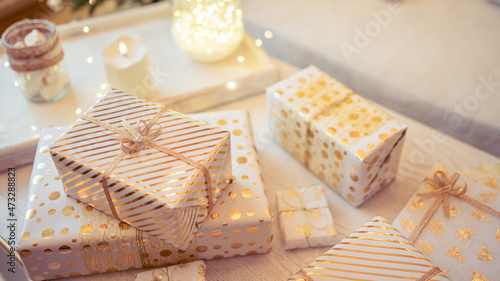 A lot of packing handmade gift boxes lying on the table near Christmas tree in the midst of golden lights, glowing garland, candle. Soft focus. 16:9
