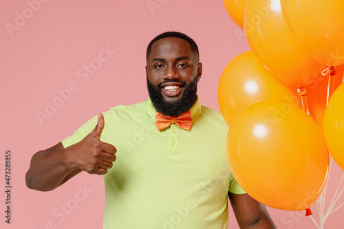 Young smiling black gay man 20s in green t-shirt hold bunch of air inflated helium balloons celebrating birthday party sow thumb up gesture isolated on plain pastel pink background studio portrait © ViDi Studio