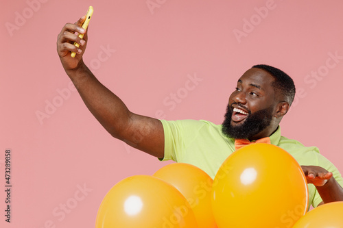 Young black gay man 20s in green t-shirt bow tie hold bunch of air inflated helium balloons celebrating birthday party doing selfie shot on mobile cell phone isolated on plain pastel pink background © ViDi Studio