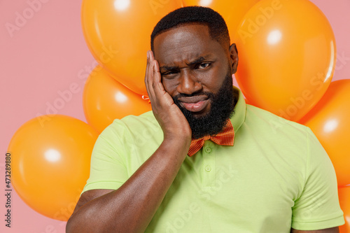 Young sad displeased black gay man in green t-shirt bow tie hold bunch of air inflated helium balloons celebrating birthday party hold face isolated on plain pastel pink background studio portrait