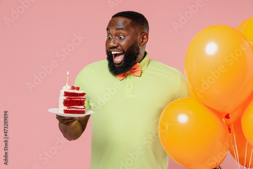 Young excited fun happy black gay man 20s in green t-shirt bow tie hold bunch of air inflated helium balloons celebrating birthday party look at sweet cake isolated on plain pastel pink background © ViDi Studio