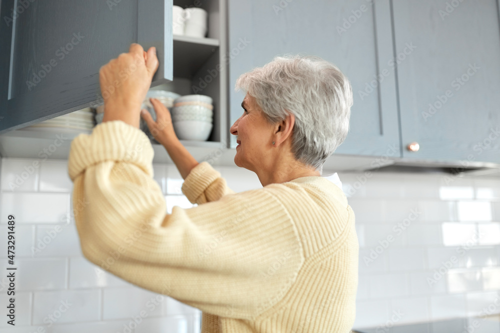 leisure, household and people concept - happy smiling senior woman opening kitchen locker with crockery at home