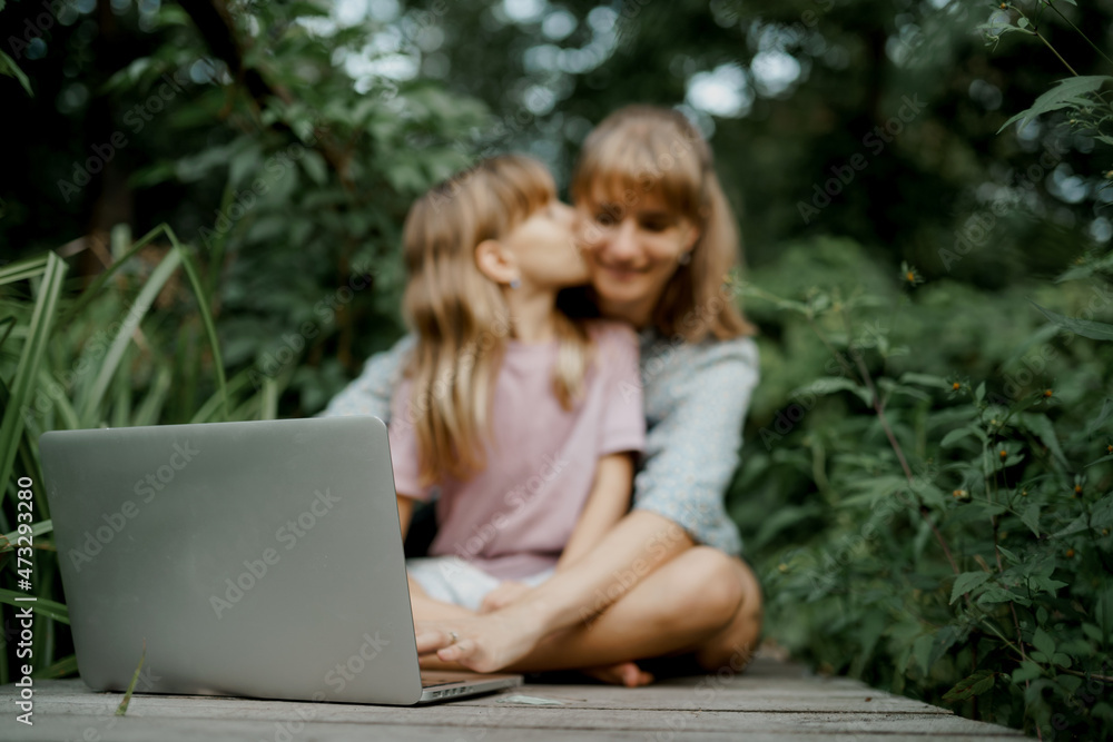 Child is kissing her mom while playing on laptop in the garden Child is kissing her mom while playing on laptop in the garden