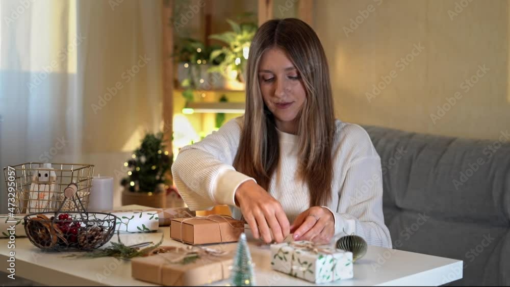 Woman wrapping Christmas eco natural gift boxes at home. Preparing presents on white table with decor elements, lights and items Christmas or New year DIY packing Concept. Woman wrapping Christmas eco natural gift boxes at home. Preparing presents on white table with decor elements, lights and items Christmas or New year DIY packing Concept.