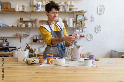 Obraz na plátně Craftswoman examining jug cap after decorating pottery with hand drawn ornament in ceramics studio or workshop