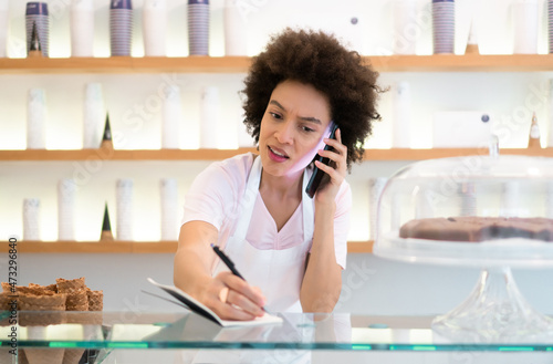 A beautiful mixed-race female worker in a pastry-gelato shop takes an order over the phone and writes it down.