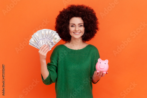Smiling affirmative woman with Afro hairstyle wearing green casual style sweater holding big sum of money and piggy bank, profitable investment. Indoor studio shot isolated on orange background.
