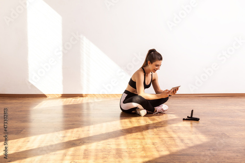 Girl sitting on floor with tablet, searching tutorial video for sport exercising, workout at home, wearing black sports top and tights. Full length studio shot illuminated by sunlight from window.