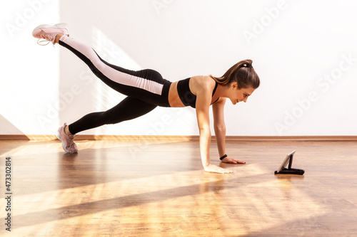 Female doing plank with raised leg, watching tutorial video on tablet, repeat after fitness blogger, wearing black sports top and tights. Full length studio shot illuminated by sunlight from window.