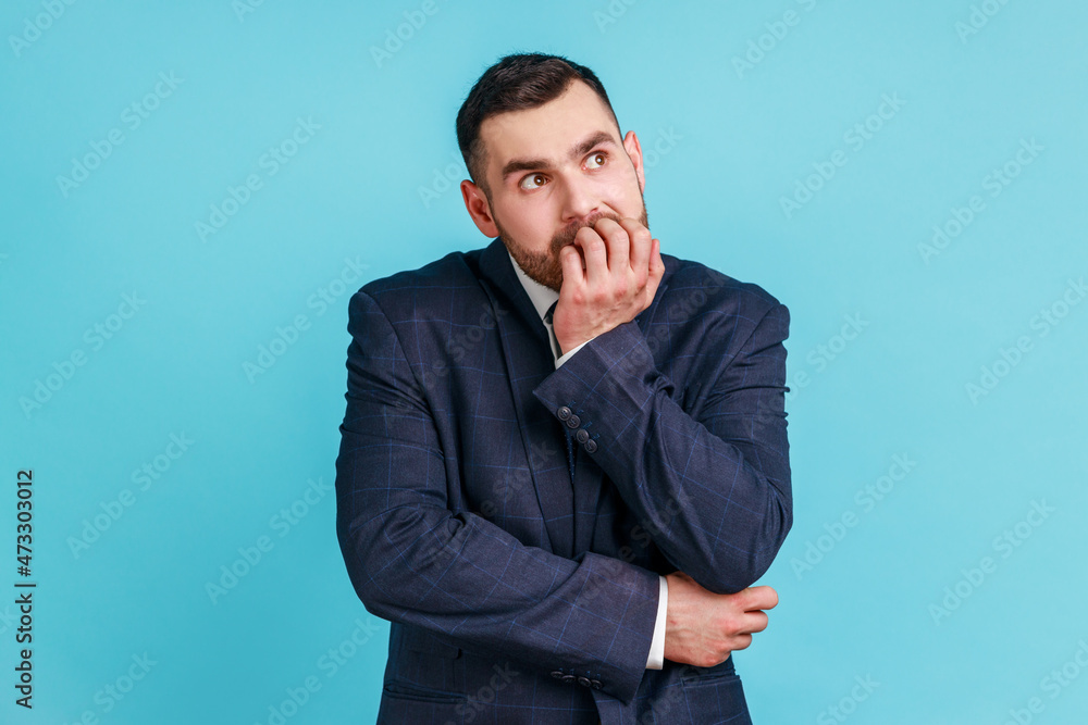 Anxious nervous businessman with beard in official style suit biting nails on fingers looking at camera with terrified expression, worried about work. Indoor studio shot isolated on blue background. Anxious nervous businessman with beard in official style suit biting nails on fingers looking at camera with terrified expression, worried about work. Indoor studio shot isolated on blue background.