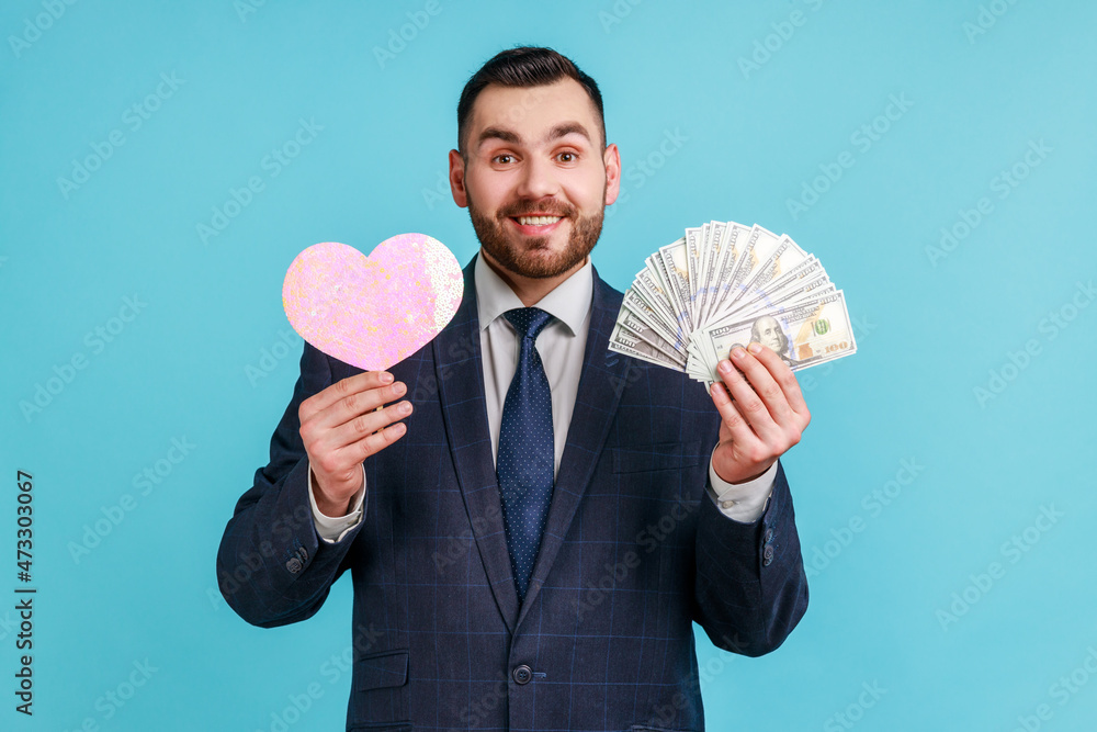 Portrait of smiling young adult bearded man wearing official style suit, holding heart and dollars banknotes, looking dissatisfied at camera. Indoor studio shot isolated on blue background. Portrait of smiling young adult bearded man wearing official style suit, holding heart and dollars banknotes, looking dissatisfied at camera. Indoor studio shot isolated on blue background.