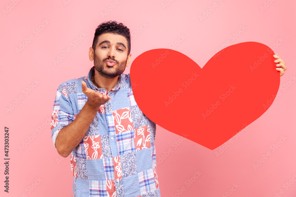 Portrait of happy romantic bearded man with big red heart in hands, looking at camera, sending air kisses, wearing blue casual style shirt. Indoor studio shot isolated on pink background. Portrait of happy romantic bearded man with big red heart in hands, looking at camera, sending air kisses, wearing blue casual style shirt. Indoor studio shot isolated on pink background.