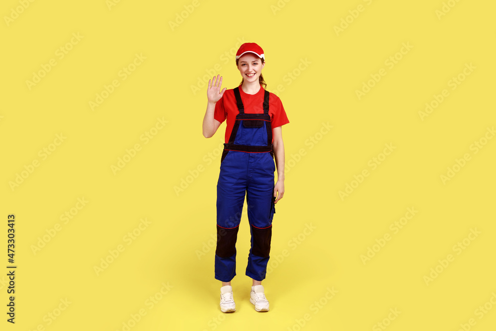 Full length portrait of smiling friendly worker woman standing and waving hand, saying hello or bye, wearing overalls and red cap. Indoor studio shot isolated on yellow background. Full length portrait of smiling friendly worker woman standing and waving hand, saying hello or bye, wearing overalls and red cap. Indoor studio shot isolated on yellow background.