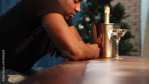 Very upset young African-American man got very drunk in a restaurant, hits the bar with his fist. Black man floods depression with alcohol on the background of a Christmas tree. Concept of a bad year