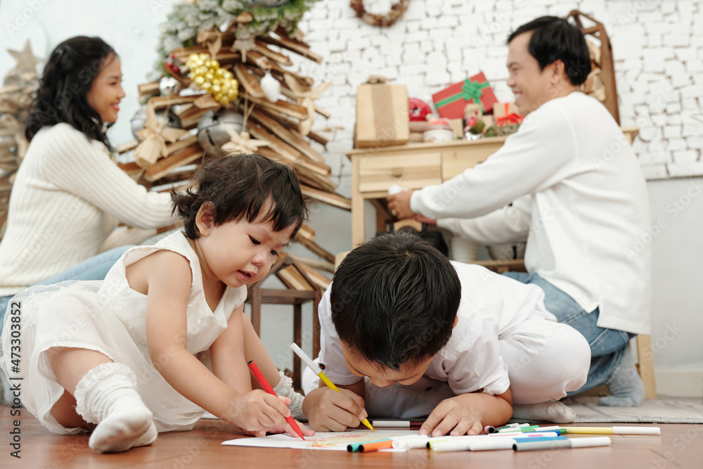 Small children drawing pictures on the floor when their parents decorating Christmas tree Small children drawing pictures on the floor when their parents decorating Christmas tree