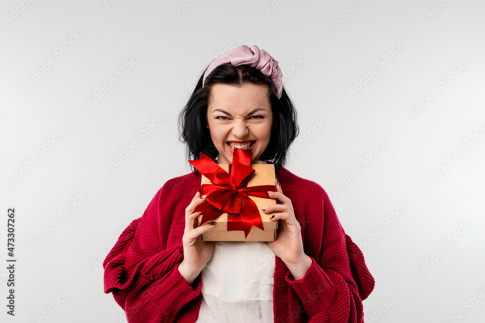 Joyful young woman in cozy red sweater holding gift box in hands biting a bow of red satin ribbon trying to unbox it, standing over white background Joyful young woman in cozy red sweater holding gift box in hands biting a bow of red satin ribbon trying to unbox it, standing over white background
