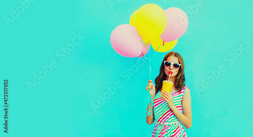 Portrait of beautiful young woman with bunch of balloons drinking a fresh juice wearing a colorful dress on blue background