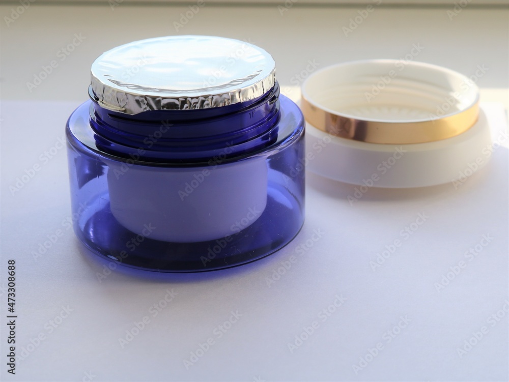 a blue plastic jar of cream with a sealed foil on top and a light lid with a gold border behind on a light background, an unused object of women's face skin care cosmetics a blue plastic jar of cream with a sealed foil on top and a light lid with a gold border behind on a light background, an unused object of women's face skin care cosmetics