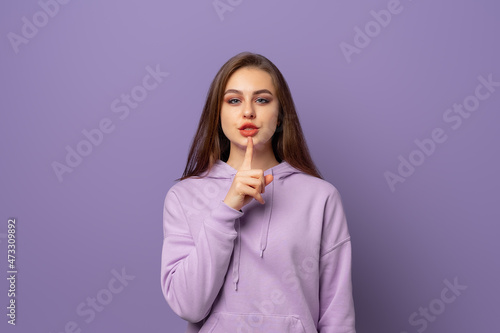Portrait of beautiful young european woman with chestnut hair holding index finger at lips, asking to keep silence or not tell anyone her secret, saying Shhh, Hush, Tsss. Keep a secret photo