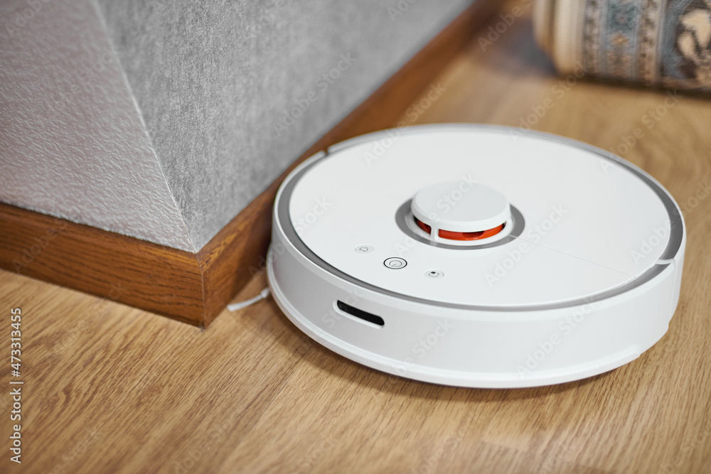 A white robot vacuum cleaner in the corner of the room. Cleaning near the baseboard. A white robot vacuum cleaner in the corner of the room. Cleaning near the baseboard.