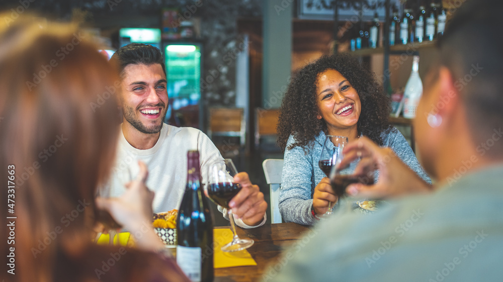 group of happy young friends having lunch at restaurant, drinking red wine and toasting, focus on the brunette colombian smiling woman, warm filter