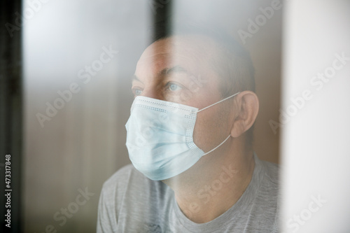 Man with a face mask sitting in a home quarantine and looking through the window.