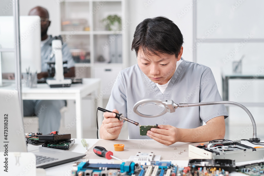 Asian woman in uniform soldering circuit board of electronic device while looking in magnifying glass Asian woman in uniform soldering circuit board of electronic device while looking in magnifying glass