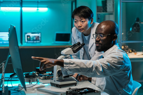 Confident African scientist pointing at computer screen while explaining something to female colleague