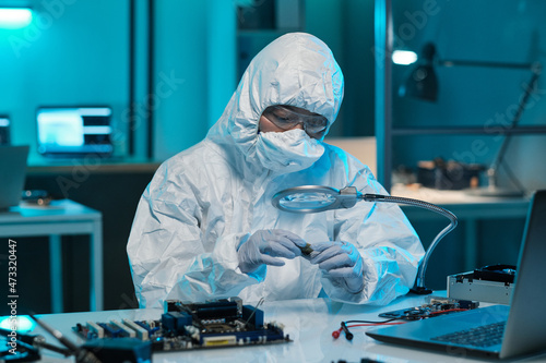 Young scientist or repairman in protective workwear looking at microchip in magnifying glass in laboratory