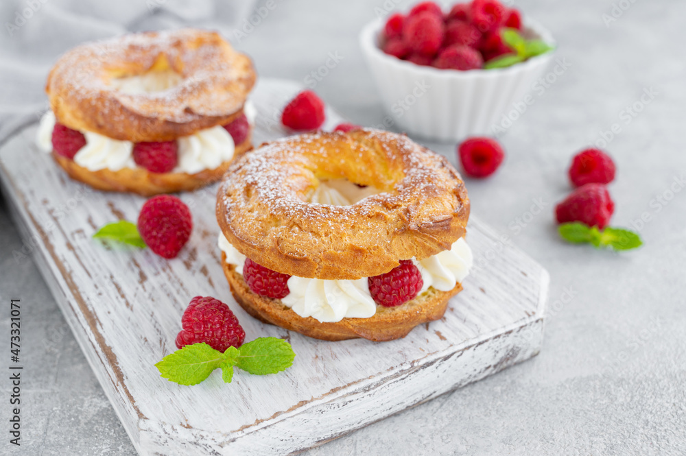 Choux pastries. Choux rings with cream of cream cheese or cottage cheese and fresh raspberries, dusted with powdered sugar on a wooden board on a gray concrete background. Choux pastries. Choux rings with cream of cream cheese or cottage cheese and fresh raspberries, dusted with powdered sugar on a wooden board on a gray concrete background.
