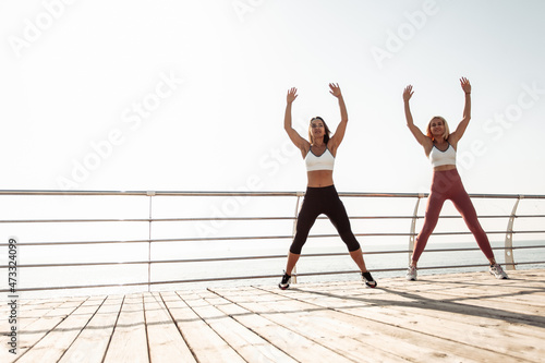 Two athletic women practicing warm-up together before training at sunrise on the beach