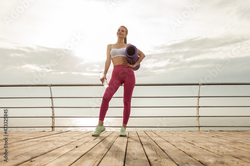 Beautiful fitness model in sportswear with water bottle and yoga mat on the beach